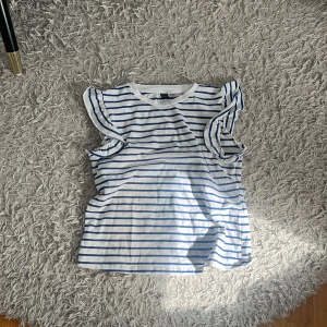 Randig volangtopp - Superfin vit topp med blå ränder från shien Modellen är ärmlös med volangdetaljer vid axlarna som ger en söt och trendig look. Tillverkad i mjuk bomull, perfekt för varma dagar. Passformen är normal och toppen är lätt att styla till jeans eller kjol.