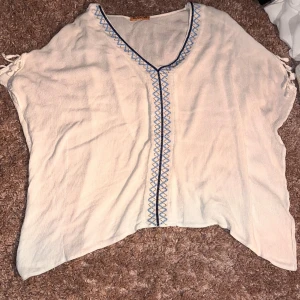 Vit blus med blå broderi från Mosnova - Vit blus från Mosnova med v-ringning och snyggt blått broderi längs halsen och mitt fram. Luftig och loose passform med knyt på axlarna. Perfekt för varma dagar och chill stil. Materialet känns lätt och mjukt mot huden.