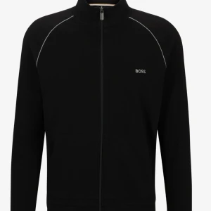 Svart zip hoodie från BOSS - Säljer en svart hoodie från BOSS med hel dragkedja. Tröjan har hög krage, långa ärmar och vita detaljer längs axlarna. Säljer den för den inte passar mig, knappt använd.