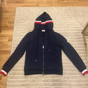 Marinblå hoodie från Moncler - Snygg marinblå hoodie från Moncler med dragkedja, huva och vita samt röda ränder på huva och ärmslut. Klassisk streetstil med fickor framtill och diskret Moncler-logga på ärmen. Perfekt för dig som gillar sportig och stilren look.