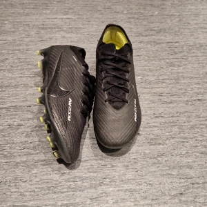 Nike Mercurial  Vapor 15 elite AG - Säljer ett par svarta Nike Air Zoom fotbollsskor med snörning och rund tå. Skorna har en snygg textur i syntetmaterial och gula detaljer på insidan och sulan. Perfekt grepp med dobbar för gräsplan. Nike-logga på sidan och AIR ZOOM-tryck.