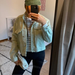 Ljusgrön croppad jeansjacka - Snygg ljusgrön jeansjacka med croppad passform och klassiska knappar framtill. Jackan har två stora bröstfickor med lock och långa ärmar. Perfekt att slänga över en t-shirt eller hoodie för en chill look.