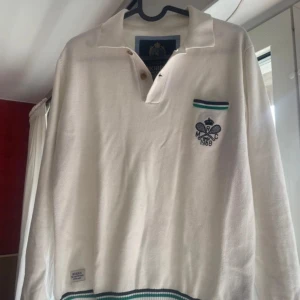 Vit långärmad pikétröja Morris - Vit pikétröja från Morris med broderad tennislogga på bröstet och randiga detaljer i grönt och svart vid krage, ficka och muddar. Tröjan har klassisk krage med två knappar och är tillverkad i mjuk bomull. Perfekt för en sportig och stilren look.