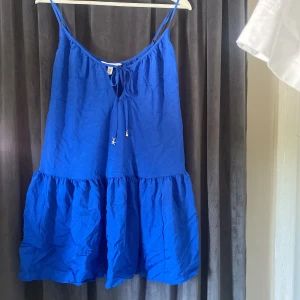 Blå kortklänning från H&M med volang - Supersöt blå kortklänning från H&M med tunna axelband och volang nertill. Klänningen har knytning framtill med små guldiga berlocker i form av sjöstjärna och snäcka. Perfekt för varma dagar och har en luftig, flowy passform.