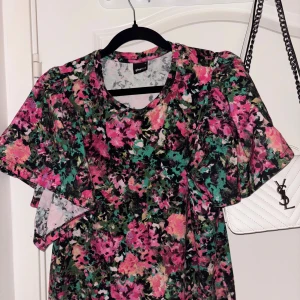 Blommig blus från Gina Tricot - Färgstark blus från Gina Tricot med blommigt mönster i rosa, grön, svart och beige. Blusen har vida korta ärmar och rund halsringning. Perfekt för dig som vill sticka ut med en lekfull och trendig look.