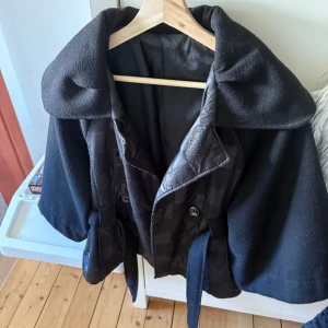 Svart kappa med bälte och stora slag - Snygg svart kappa med breda slag och bälte i midjan. Jackan har rutigt mönster i olika material på fram- och baksidan, vilket ger en cool kontrast mellan blankt och matt tyg. Perfekt för dig som vill sticka ut med en unik och trendig look.