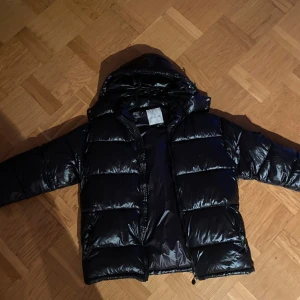 Svart Moncler Maya dunjacka  - Snygg svart dunjacka från Moncler med glansig finish och klassisk logga på ärmen. Jackan har huva, dragkedja framtill och praktiska fickor med dragkedja. Perfekt för kalla dagar när du vill hålla stilen och värmen.