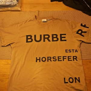 Beige Burberry t-shirt med tryck - Snygg beige t-shirt från Burberry med svart grafiskt tryck över hela framsidan och ena ärmen. Klassisk rund halsringning och korta ärmar. Tillverkad i 100% bomull för en skön och mjuk känsla. Perfekt för dig som gillar streetstyle och designerplagg.