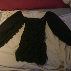 Svart kort klänning med puffärmar - Supersnygg svart kort klänning med markerad rynkning och stora puffärmar. Klänningen har en tight passform över kroppen och vida ärmar som ger en cool statement-look. Perfekt för dig som vill sticka ut och känna dig extra trendig.