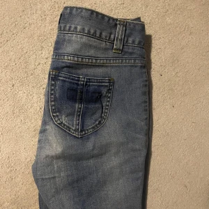 Blå jeans med fickdetaljer - Snygga blå jeans med slitningar och coola fickor med lock och knapp bak. Jeansen har en klassisk femficksdesign, bälteshällor och ljus tvätt för en avslappnad look. Perfekta för dig som gillar streetstyle och vill ha något unikt i garderoben. Strl 38 