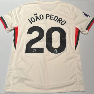 Chelsea João Pedro #20 bortatröja Nike - Chelsea FC bortatröja med João Pedro och nummer 20. Tröjan är vit med tunna röda och mörkblå ränder framtill, broderad Nike-logga och Chelsea-märke på bröstet. Premier League-logga på ärmen. Tillverkad i lätt och ventilerande polyester. Replika, endast handtvätt gäller!