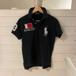 Svart Polo Ralph Lauren Bahrain t-shirt - Svart t-shirt från Polo Ralph Lauren med Bahrain-flagga och text på både fram- och baksida. Stor vit polospelare broderad på bröstet och siffran 3 på ärmen och ryggen. Klassisk pikékrage och korta ärmar. Perfekt för dig som gillar sportig och stilren look.