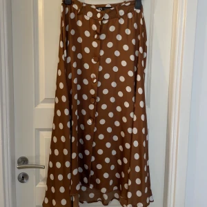 Brun prickig midikjol från Zara - Superfin brun kjol från Zara med stora vita prickar och knappar framtill. Kjolen har en lätt A-linjeformad siluett och är tillverkad i ett mjukt, glansigt material som ger ett snyggt fall. Perfekt för dig som gillar retrovibbar och vill sticka ut lite extra.