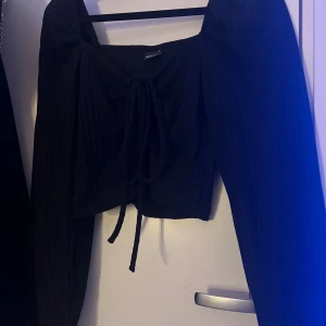 Svart croppad topp med knyt - Säljer en svart croppad topp med långa ärmar och snygg knytning framtill. Toppen har en fyrkantig urringning och är ribbad för extra stilpoäng. Perfekt att matcha med höga jeans eller kjol.