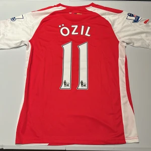 Arsenal Özil 11 hemmatröja Puma - Röd och vit Arsenal hemmatröja från Puma med Özil och nummer 11 på ryggen. Premier League-patch på ärmarna och Fly Emirates-tryck på bröstet. Tillverkad i lätt och ventilerande material, perfekt för fotboll eller samling. Replika, endast handtvätt gäller!
