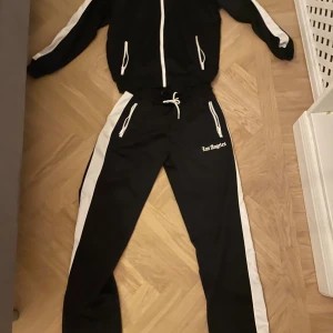 Svart tracksuit från new yorker - Svart tracksuit med vita sidoränder och snörning i midjan. Väldigt bra kvalitet. Byxorna och tröjan säljs som ett paket!