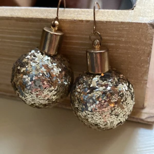Glittriga guldiga örhängen - Säljer ett par unika örhängen formade som små julkulor, täckta av glittrigt guldigt glitter. De har en rund form och metallfäste i guldton. Perfekta för dig som vill sticka ut och addera extra bling till din stil.
