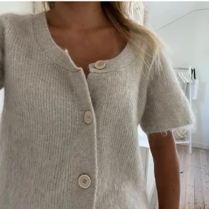 Beige stickad kofta med kort ärm - Supermjuk och fluffig beige stickad kofta med korta ärmar och stora knappar framtill. Perfekt för lager-på-lager och har en avslappnad, lite boxig passform. Enkel att styla till både jeans och kjol.