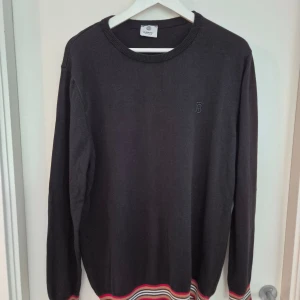 Svart Burberry tröja med ränder - Svart långärmad tröja från Burberry i 100% bomull. Tröjan har rund halsringning och diskret broderad logga på bröstet. Färgglada ribbade muddar i rött, beige, vitt och svart vid ärmslut och nederkant ger en snygg kontrast.