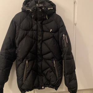 Svart dunjacka Peak Performance S - Svart dunjacka från Peak Performance med huva och dragkedja. Jackan har quiltad design, två fickor med dragkedja framtill och en ärmficka. Perfekt för kalla dagar och har en sportig vibe. Snygg passform och klassisk look. Köpt för 2 900 kr.