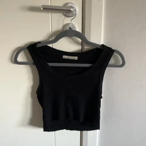 Svart ribbad croppad linnetopp - Svart croppad linnetopp från Pull&Bear med breda axelband och rundad halsringning. Toppen är ribbad och har en tight passform, perfekt att matcha med jeans eller kjol. Enkel och stilren design som funkar till många olika looks.