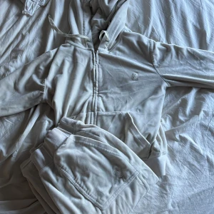 Juicy couture  - Mysigt vitt sett från Juicy Couture i mjuk velour med dragkedja framtill, huva med snörning och broderad logga på bröstet. Perfekt för en avslappnad och trendig look. Byxorna saknar snöre