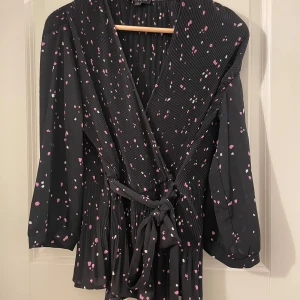 Svart omlottklänning Selected Femme - Svart kort omlottklänning från Selected Femme med plisserat tyg och knyt i midjan. Klänningen har lång ärm och är täckt av små rosa, vita och blå prickar. Perfekt för dig som gillar en lekfull men stilren look.