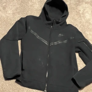 Svart Nike hoodie med dragkedja - Svart hoodie från Nike med hel dragkedja och huva. Snygga svarta detaljer i form av ett diskret mönster över bröstet och Nike-logga. Tröjan har långa ärmar och är gjord i mjuk bomullsmix, perfekt för chill eller träning.    Nike tech fleece Svart Använd och har defekter som ni ser på bilden  Har INTE försökt att tvätta bort men kanske går bort under tvätt om inte kan ni nog bara skrubba lätt bort.  Hål som man också ser men är nog inte så sort problem(fler små små hål men syns knapp)