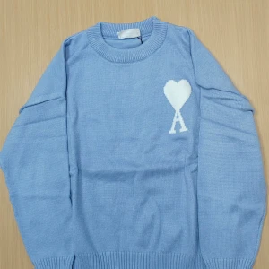Blå stickad Ami tröja - Ljusblå stickad tröja från AMI Paris med vit hjärtdetalj och bokstaven A på bröstet. Tröjan har rund halsringning, ribbade muddar och lång ärm. Perfekt för dig som gillar stilrena och trendiga plagg med en twist.
