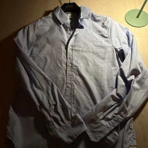 Ljusblå skjorta från BOSS, strl S - Säljer en stilren ljusblå skjorta från BOSS i storlek S. Skjortan har klassisk krage, knäppning framtill och broderad BOSS-logga på bröstet. Tillverkad i mjuk bomull och har långa ärmar med manschett. Perfekt för en clean och snygg look.