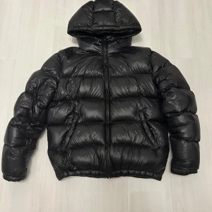 Svart pufferjacka från Zara med huva - Säljer en svart pufferjacka från Zara i storlek M. Jackan har en rejäl huva, dragkedja framtill och en glansig finish. Perfekt för kalla dagar när du vill hålla värmen och ändå se stilren ut.