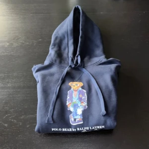 Helt ny Ralph Lauren hoodie  - Säljer en mörkblå hoodie från Polo Ralph Lauren med ikoniska Polo Bear-trycket framtill. Tröjan har huva med snörning och är tillverkad i 100% bomull för en skön och avslappnad känsla. Perfekt för dig som gillar klassisk streetstyle.