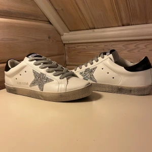 Golden Goose vita sneakers med glitterstjärna - Golden Goose sneakers i vitt skinn med grå snörning och svart häl. Sidan pryds av en glittrig silverstjärna som ger extra edge. Sulan har en vintage-look och skon har rund tå och platt sula. Perfekta för dig som vill sticka ut med stil.