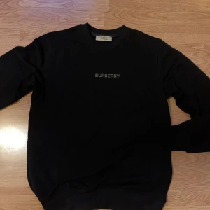 Svart sweatshirt från Burberry - En stilren svart sweatshirt från Burberry med diskret logga på bröstet. Klassisk rund halsringning och långa ärmar. Perfekt för dig som gillar enkel men exklusiv stil. Tillverkad i mjuk bomull för maximal komfort.
