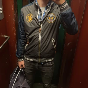 Jacka från saint Monica  - Jacka från Saint Monica  med broderade patchar på bröstet och ärmen. Jackan har dragkedja, ribbade muddar och detaljer i vitt och grått. Perfekt för dig som vill sticka ut med en sportig och cool stil.