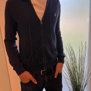 Mörkblå stickad hoodie från Ralph Lauren - Säljer en mörkblå stickad hoodie från Ralph Lauren med dragkedja och huva. Tröjan har snörning vid huvan, ribbade muddar och två fickor framtill. Klassisk logga broderad på bröstet. Perfekt för dig som gillar en chill och stilren look.