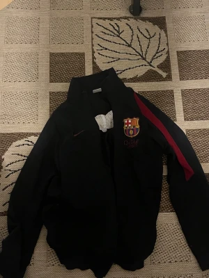 Svart FC Barcelona vindjacka Nike - Svart vindjacka från Nike med FC Barcelona-logga på bröstet och röd detalj längs ärmarna. Jackan har hög krage, dragkedja och texten 'BARÇA' på ryggen. Perfekt för dig som älskar fotboll och vill visa ditt stöd för laget. Storlek xs 