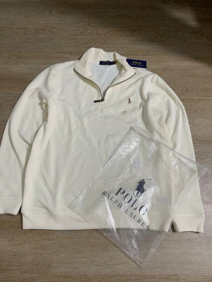 Vit halvzip från Polo Ralph Lauren - Klassisk vit halvzip-tröja från Polo Ralph Lauren med broderad logga på bröstet. Tröjan har hög krage med dragkedja och ribbade muddar vid ärmslut och nederkant. Perfekt för en clean och stilren look.