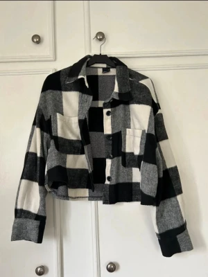 Svartvit rutig overshirt - Trendig overshirt i svartvitt rutigt mönster med stora rutor. Modellen är croppad med klassisk krage, knäppning framtill och två bröstfickor. Materialet ser ut att vara mjukt och lite tjockare, perfekt för lager-på-lager. Overshirten har långa ärmar med breda muddar.