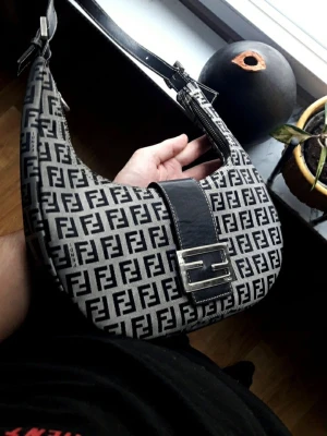 Fendi Baguette monogram handväska - Säljer en Fendi Baguette handväska i svart och beige med det klassiska FF-monogrammet över hela väskan. Finns lite slitage men annars jätte bra skick som en gåva eller present till någon, bra utrymme och finns även en liten extra fack i väskan,Väskan har också ett justerbar axelrem i svart läder.