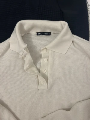 Vit långärmad skjorta från Zara - Klassisk vit långärmad skjorta från Zara med krage och knappar framtill. Skjortan är i ett mjukt, stickat material och har en enkel, stilren design som passar till många olika looks. OBS! Tröjan går ner till naveln på mig och jag är 170. Det är en topp basically 