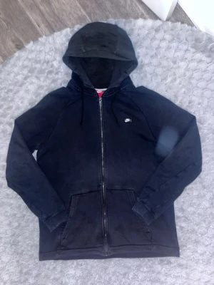 Svart zip hoodie från Nike - Säljer en svart hoodie från Nike med dragkedja framtill och klassisk huva med snören. Hoodien har två stora fickor på magen och en liten Nike-logga på bröstet. Tillverkad i mjukt bomullsmaterial, perfekt för chill dagar eller när du vill ha en clean streetstyle.