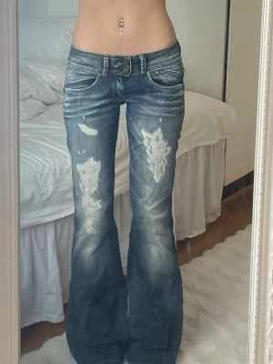 Blå bootcut jeans med slitningar/hål - Snygga blå bootcut jeans med slitna detaljer och dubbla knappar i midjan. Jeansen har en låg midja och utställda ben, perfekt för en trendig och avslappnad look. Storlekslappen har suddats ut men uppskattar XS-S 🩷