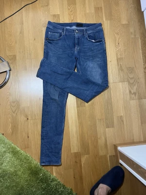 Blå slim fit jeans från Tiger of Sweden - Snygga blå slim fit jeans från Tiger of Sweden med klassisk femficksdesign och silverfärgad knapp. Jeansen har normal midja och smala ben, perfekta för en stilren look. Materialet är klassisk denim i bomull. Strl 33/32