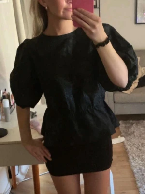 Svart puffärmad kort klänning - Säljer en svart blus med puffiga korta ärmar och rund halsringning. Blusen har en lätt glansig yta och markerad midja som ger en snygg siluett. Perfekt för dig som vill ha en trendig och stilren topp.💗💗
