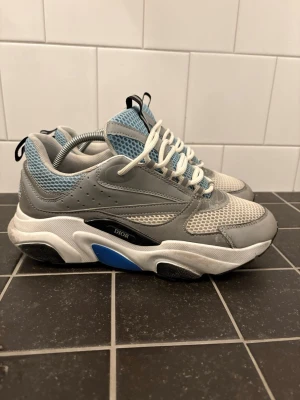Dior B22 sneakers grå/blå/vit - Chunky Dior B22 sneakers i grått, blått och vitt med meshpaneler, läderdetaljer och tjock sula. Skorna har vita snören, svart och blå detalj på sulan samt svart dragflik på plösen. Perfekt för dig som gillar statement sneakers och streetwear.