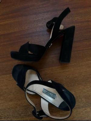 Svarta klackar från Prada - Snygga svarta pumps från Prada med bred klack och platåsula. Skorna har en öppen tå, korsade remmar över foten och justerbar ankelrem. Tillverkade i mocka för en lyxig känsla och passar perfekt till dig som vill ha både höjd och stabilitet.