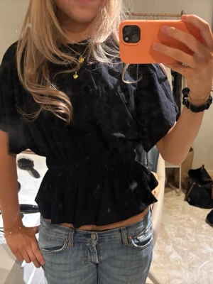 Svart volangblus med resår i midjan - Trendig svart topp med volangkant och resår i midjan som ger en snygg siluett. Modellen är croppad och har korta ärmar. Perfekt att matcha med jeans eller kjol för en cool look. Använd 1 gång