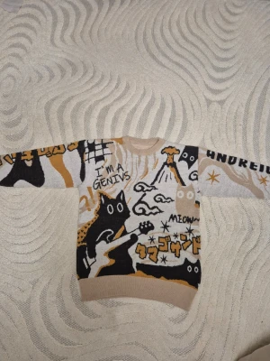 Stickad tröja med kattmotiv och text - Cool stickad tröja i beige, svart, vitt och senapsgult med grafiskt kattmotiv, texten 'I'M A GENIUS' och japanska tecken. Tröjan har rund hals, ribbade muddar och unikt mönster med stjärnor och vulkan. Perfekt för dig som gillar statement-plagg.
