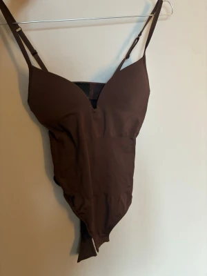 Brun body med smala axelband - Säljer en brun body med smala, justerbara axelband och v-ringad urringning. Bodyn är figurnära och har en enkel, stilren design i stretchigt material. Perfekt att styla med jeans eller kjol för en trendig look. Endast använd 1 gång 